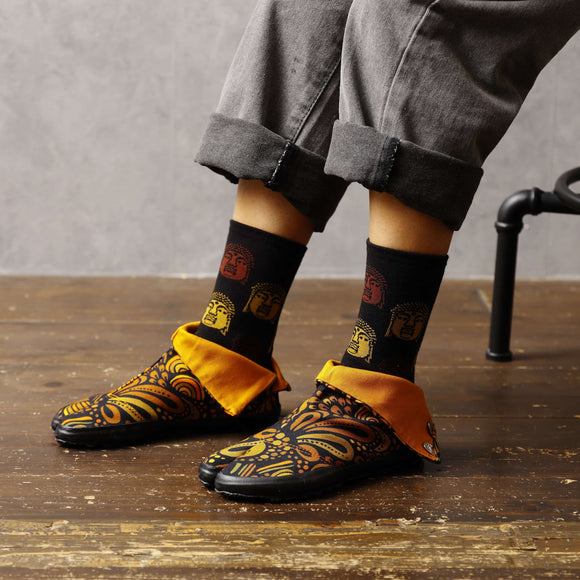 "Side view of a person wearing UROKO Tabi Shoes in orange, paired with black socks featuring colorful Daruma designs and rolled-up gray pants."